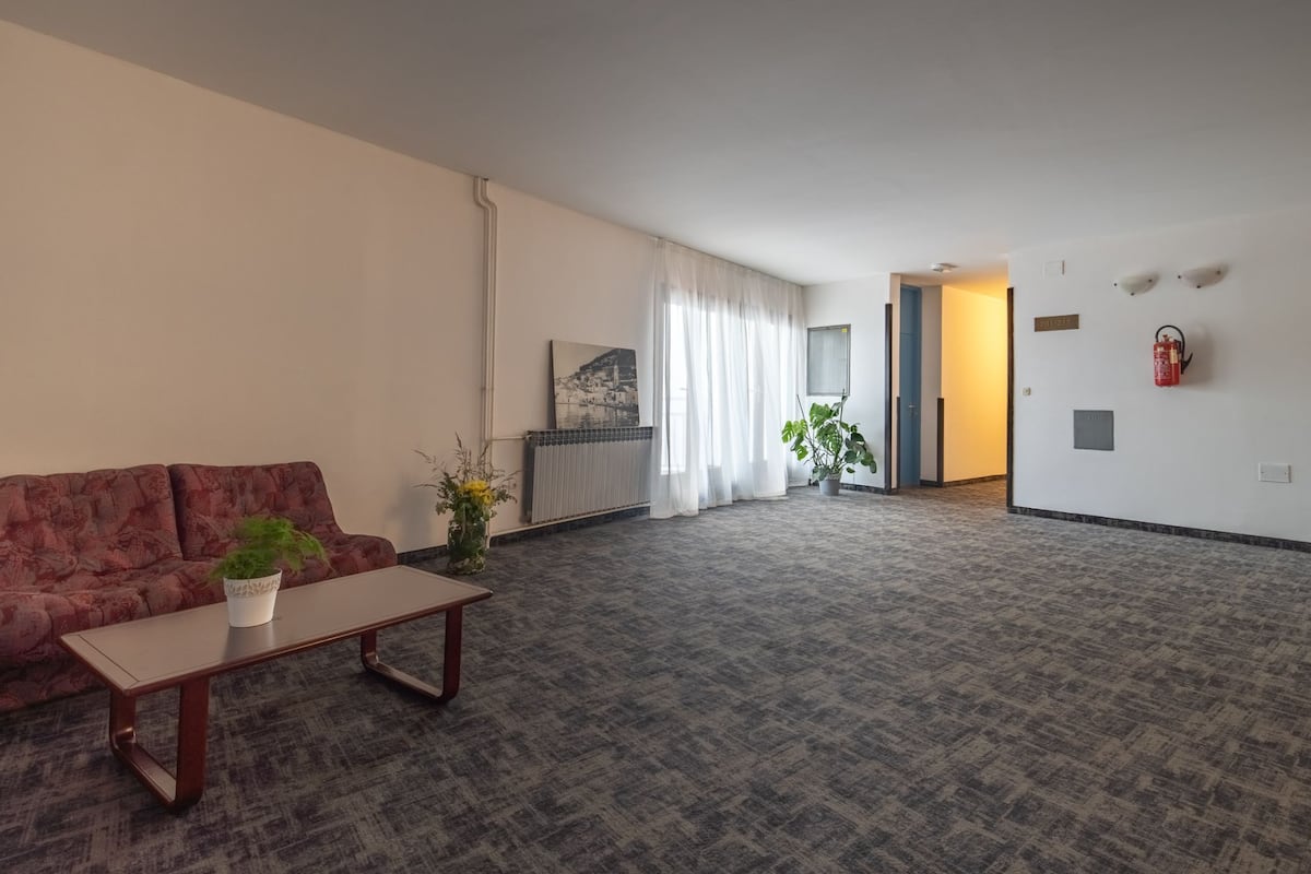Uma sala de estar espaçosa com sofá, mesa de centro e um vaso de planta. Há um quadro na parede e um extintor fixado na parede.