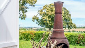 Een rustieke buitenomgeving met een grote metalen schoorsteen, een rieten mand vol takken, en een wit gebouw op de achtergrond.