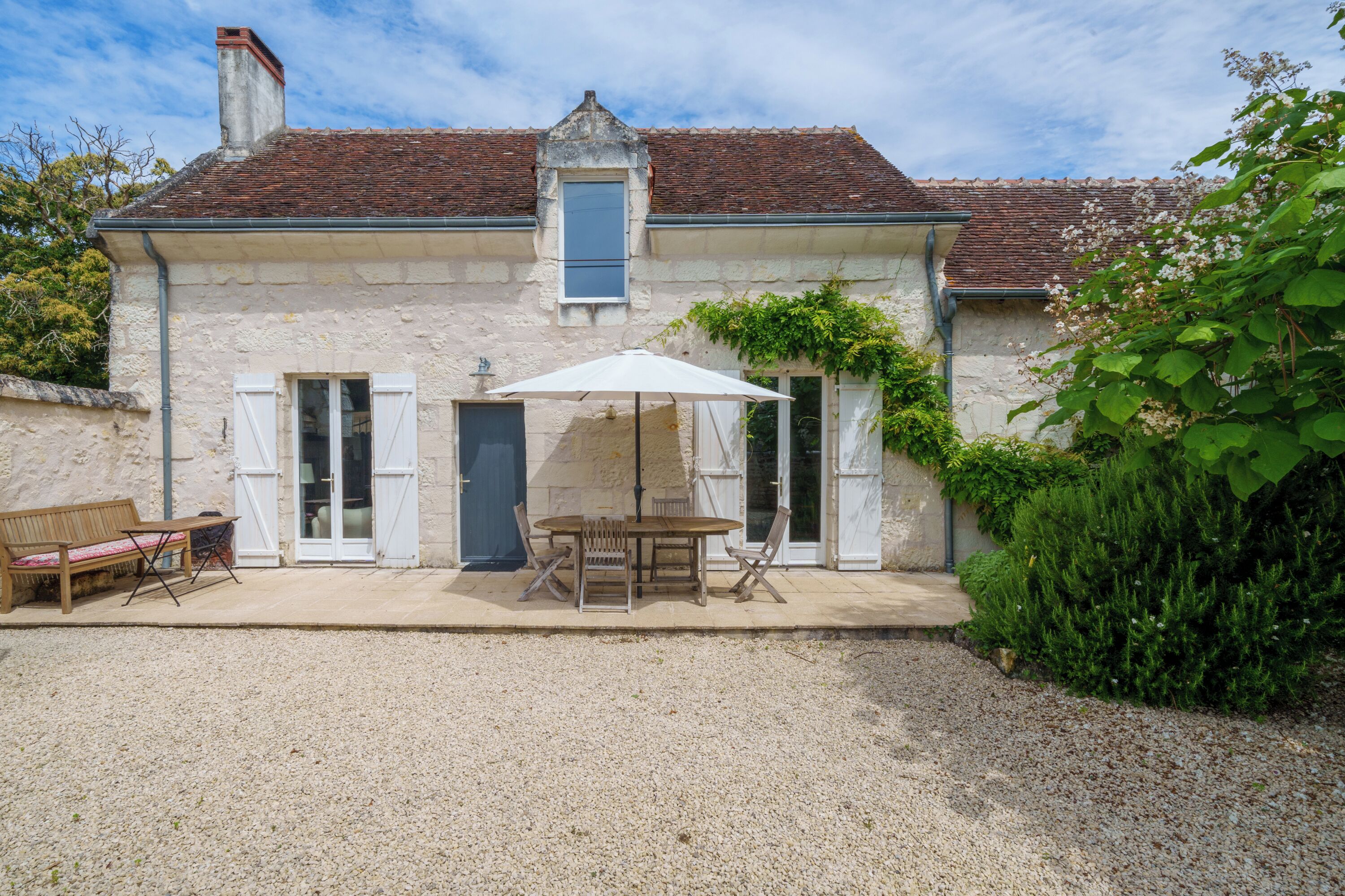 Un bâtiment en pierre avec un toit en tuiles, un patio couvert avec une table et des chaises, et un sol en gravier.