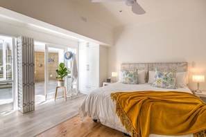 Une chambre à coucher comprenant un lit, des tables de chevet, une petite table, un ventilateur de plafond, une plante en pot et une porte-fenêtre coulissante donnant sur un espace extérieur.