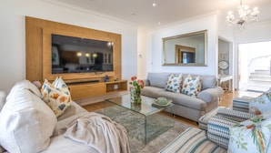 A living room with a large flat-screen TV, a grey sofa with floral cushions, a glass coffee table, and a patterned rug. - Villa Rice - Eight Bedroom Villa, Sleeps 16 (Plettenberg Bay)