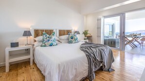 A modern bedroom with a large bed, two nightstands, a bedside table, and a view of an outdoor balcony. - Villa Rice - Eight Bedroom Villa, Sleeps 16 (Plettenberg Bay)