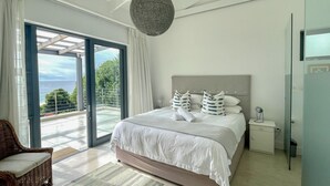 A bedroom with a large bed, a view of the outdoors, and a wicker chair. - Estrella Residence - Four Bedroom Villa, Sleeps 12 (Plettenberg Bay)