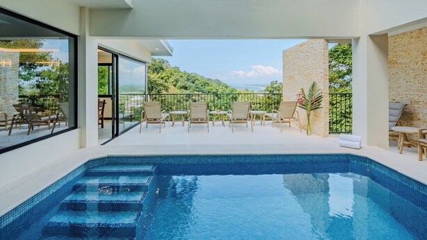 A modern indoor swimming pool with clear blue water, surrounded by wooden lounge chairs and a glass balustrade.