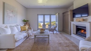A modern living room with a white sofa, a coffee table, and a fireplace. There is a large window with a view of the ocean.