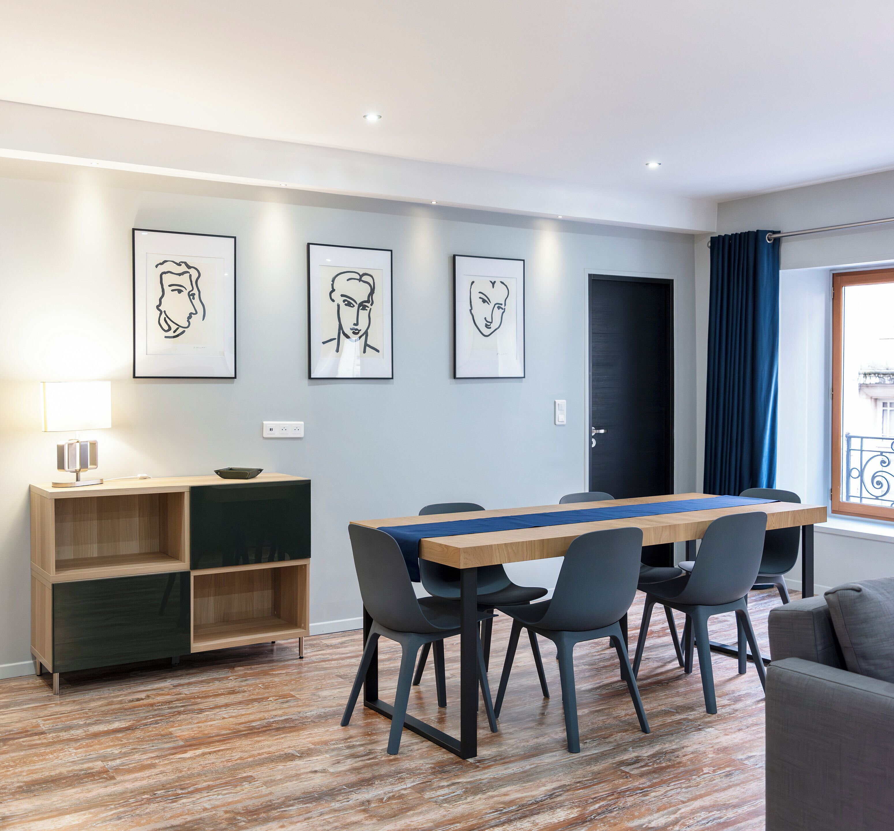 A modern living room with a wooden dining table, grey chairs, a grey sofa, and a blue curtain. There are three framed artworks on the wall.