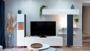A modern living room with a flat-screen TV, white cabinetry, and a small dining area with two stools. - Rock in Share - Les Ponts (Nancy)