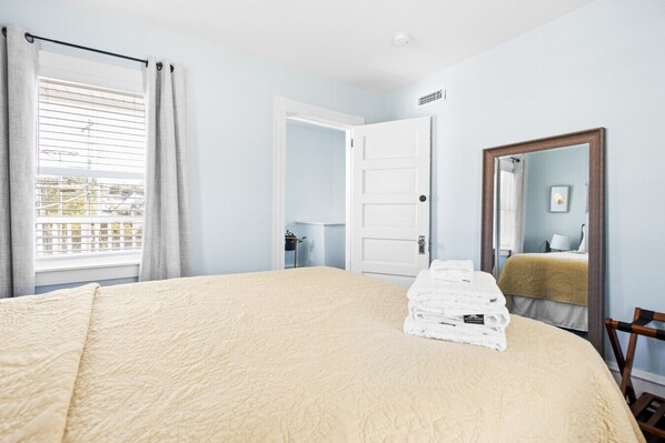 A hotel room with a bed, a stack of towels, a window with blinds, a door, and a mirror.