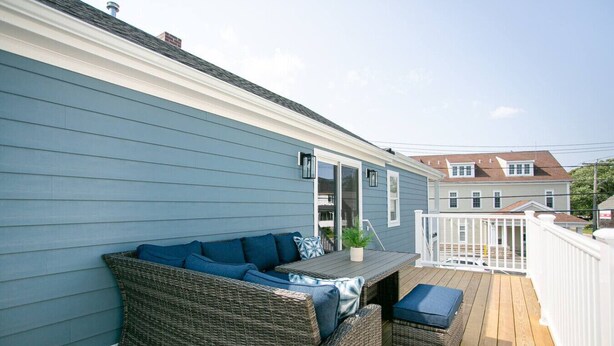 A covered outdoor deck with a wicker sofa and a small table, a white railing, and a view of neighboring houses.