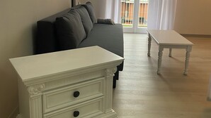 Et værelse med en grå sofa, et hvidt natbord med skuffer, et lille hvidt bord og en balkon med hvide gardiner.