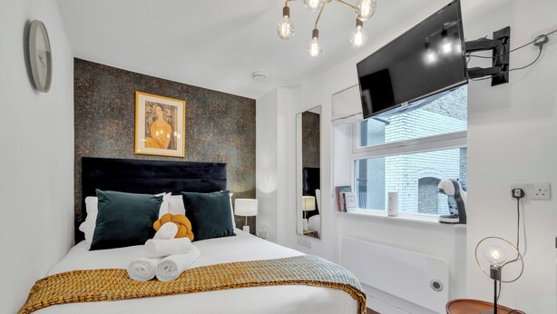 A modern hotel room with a large bed, a mounted TV, a window with a view, and a unique light fixture. - Cozy Studio in Central Locale with Kitchenette (London)