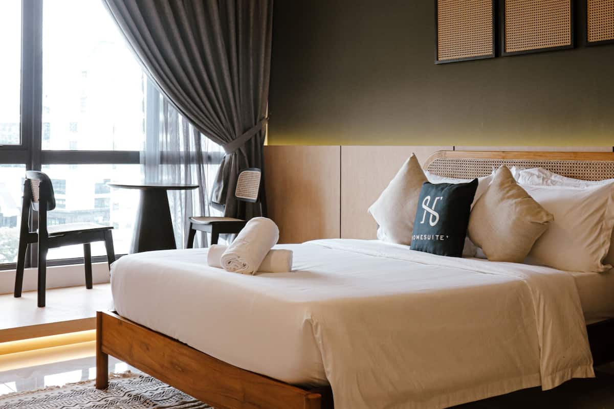 Cama arrumada com almofadas e uma toalha enrolada, uma mesinha com uma cadeira, e vista da cidade através de grandes janelas.