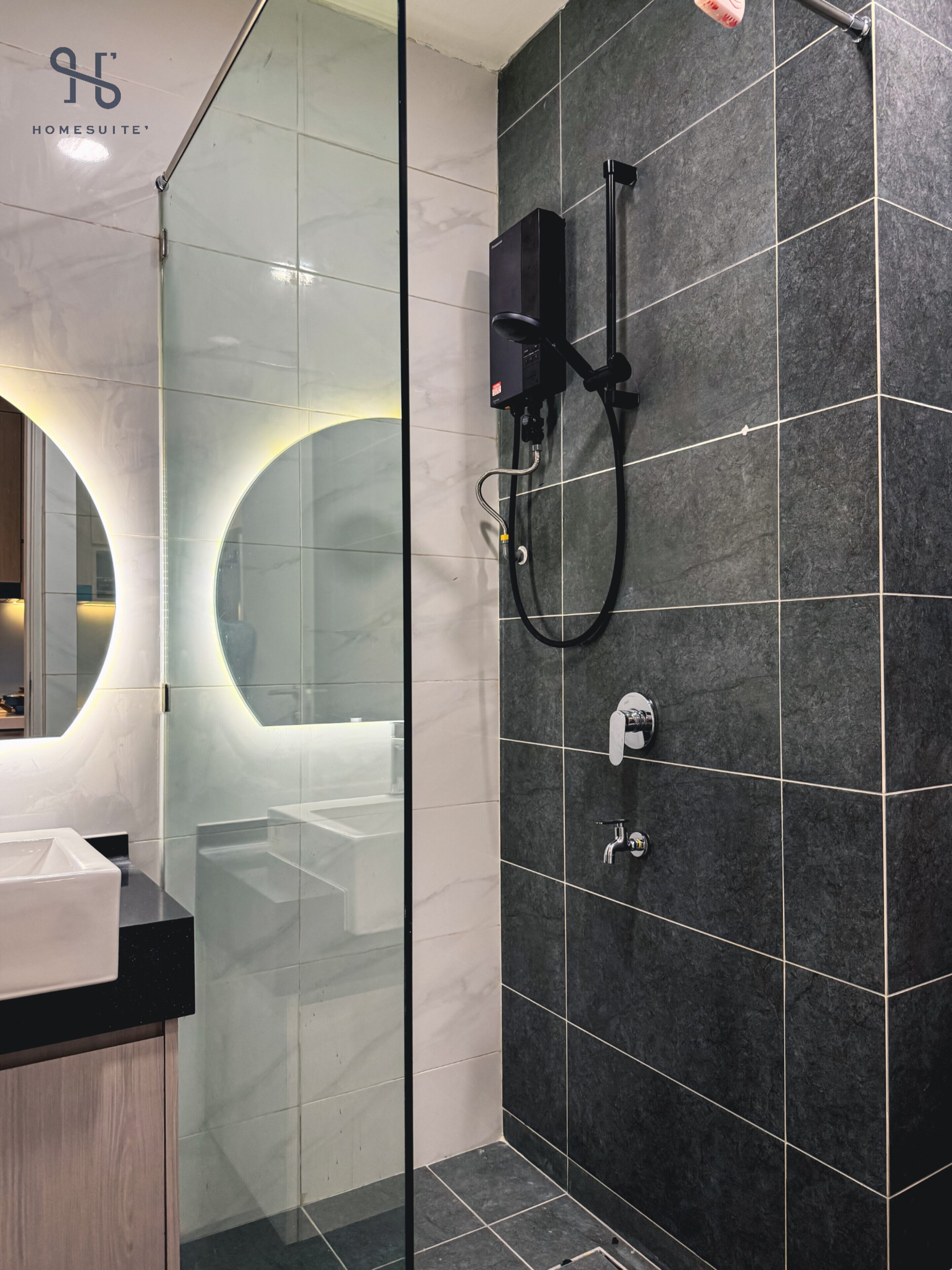 A modern bathroom with a glass shower enclosure, a large mirror, and a dark tiled wall.