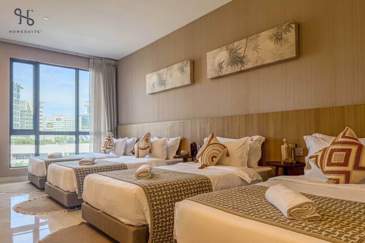 Quarto de hotel com três camas, janelas amplas e obras de arte nas paredes.