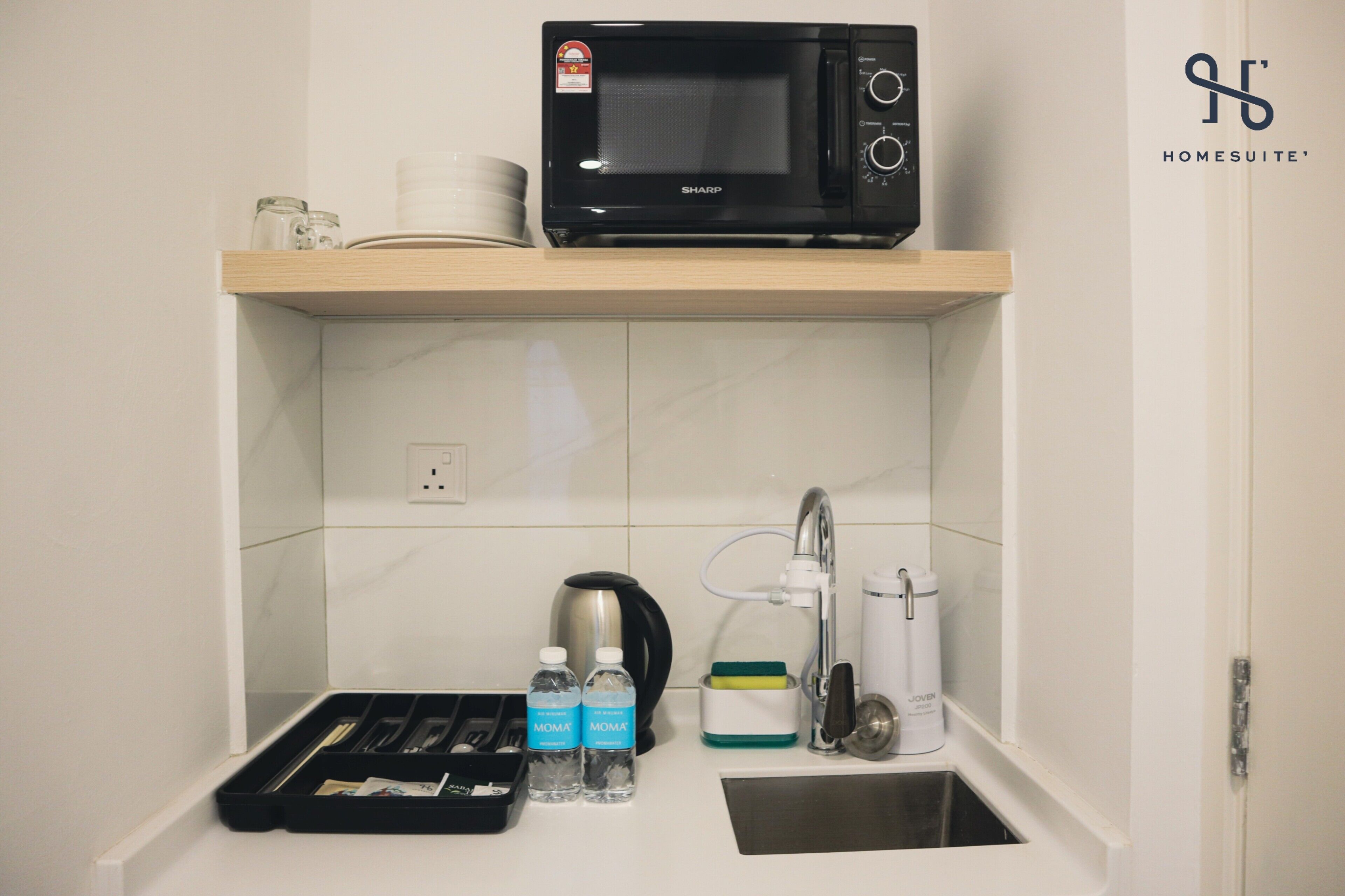 A compact kitchen with a microwave on a shelf, a kettle, a coffee maker, and a tray with utensils.