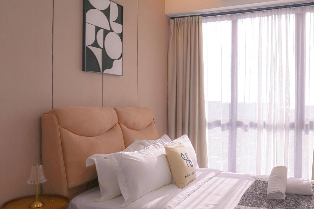 Quarto de hotel com uma cama grande, cabeceira, mesa de cabeceira com abajur, janela com cortinas e ar condicionado.