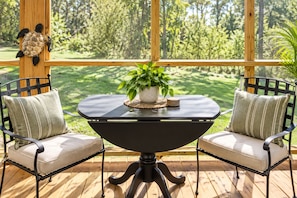 Een houten terras met een ronde tafel en twee stoelen, een bloempot op de tafel, en een uitzicht op een groen landschap door grote ramen.