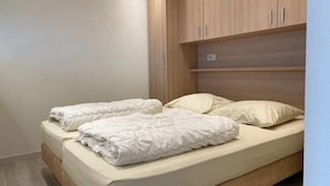 Una cama individual con colchón color crema y ropa de cama blanca, cabecera de madera y una pequeña estantería arriba.