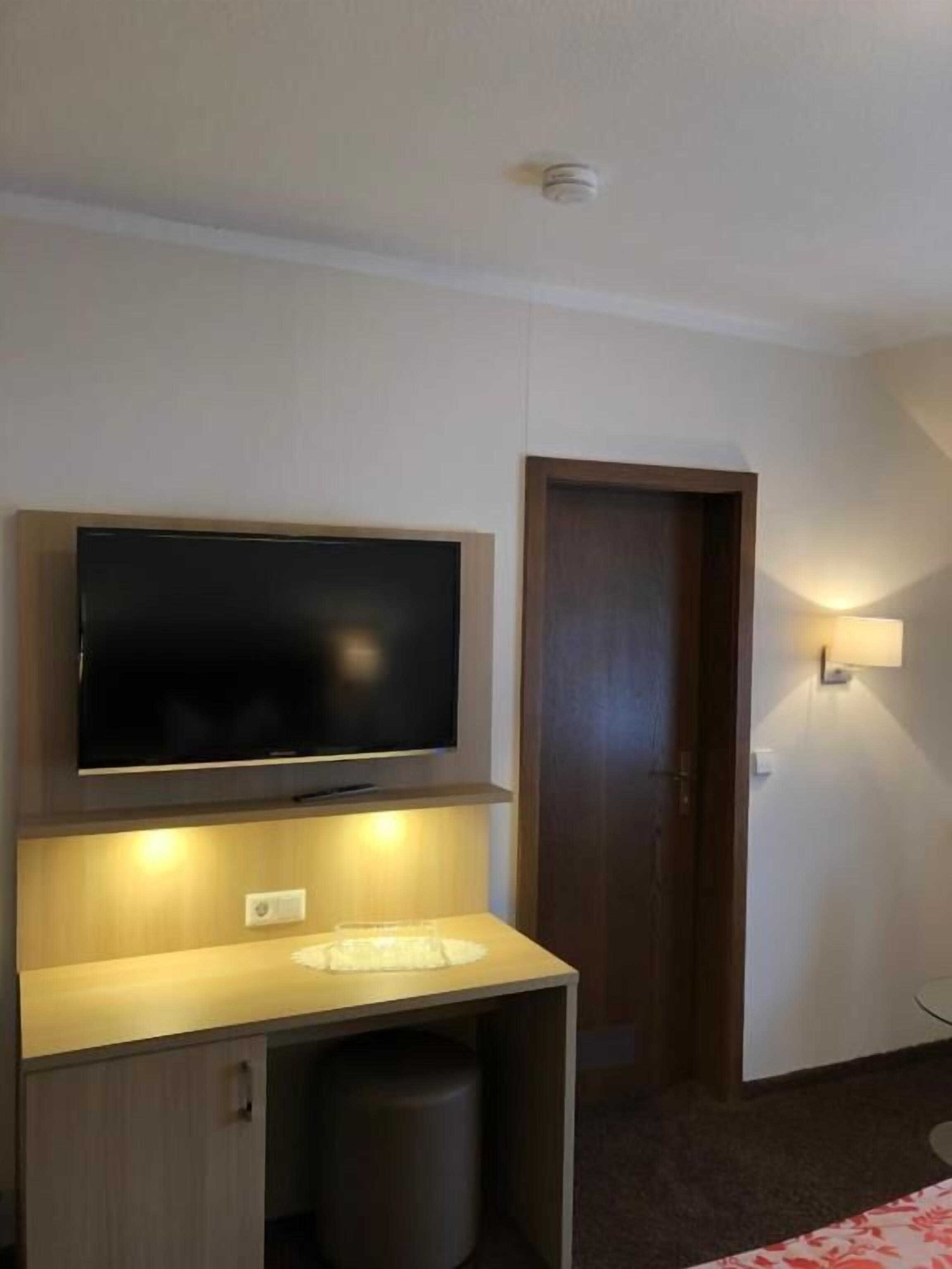 Ein Hotelzimmer mit einem an einer Holzkassette befestigten Flachbildfernseher, einer kleinen Mülltonne, einer Tür und einer wandmontierten Leuchte.