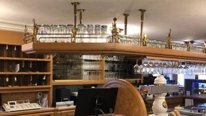 Zona de bar con estantes de madera que sostienen vasos y botellas, una caja registradora y un monitor de computadora.