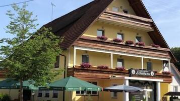 Un edificio a due piani con tetto spiovente, balconi in legno con fioriere e un cartello con su scritto "Schulzen".