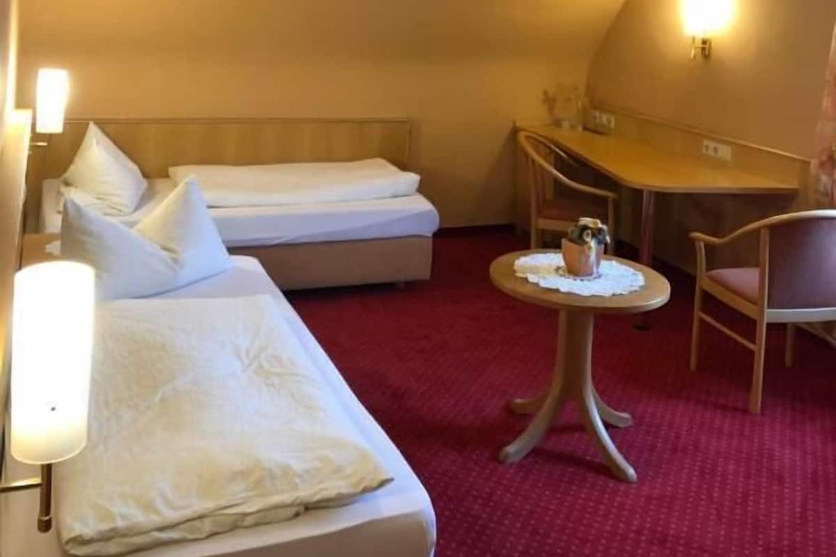 A hotel room with two beds, a small round table with a lamp, a chair, and a desk.