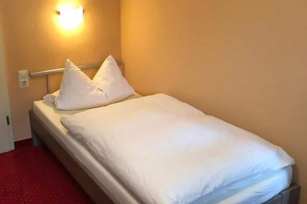 A single bed with white bedding and pillows, a wall-mounted light, and a red carpeted floor.