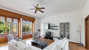 A spacious living room with a white sofa, a black coffee table, and a television. There are wooden cabinets, a floor lamp, and a ceiling fan. - Escape to Apulia by BarbarHouse (Pulsano)
