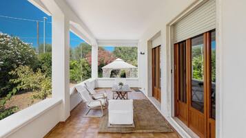 A covered patio with a wooden floor, white furniture, and a view of greenery and a gazebo.