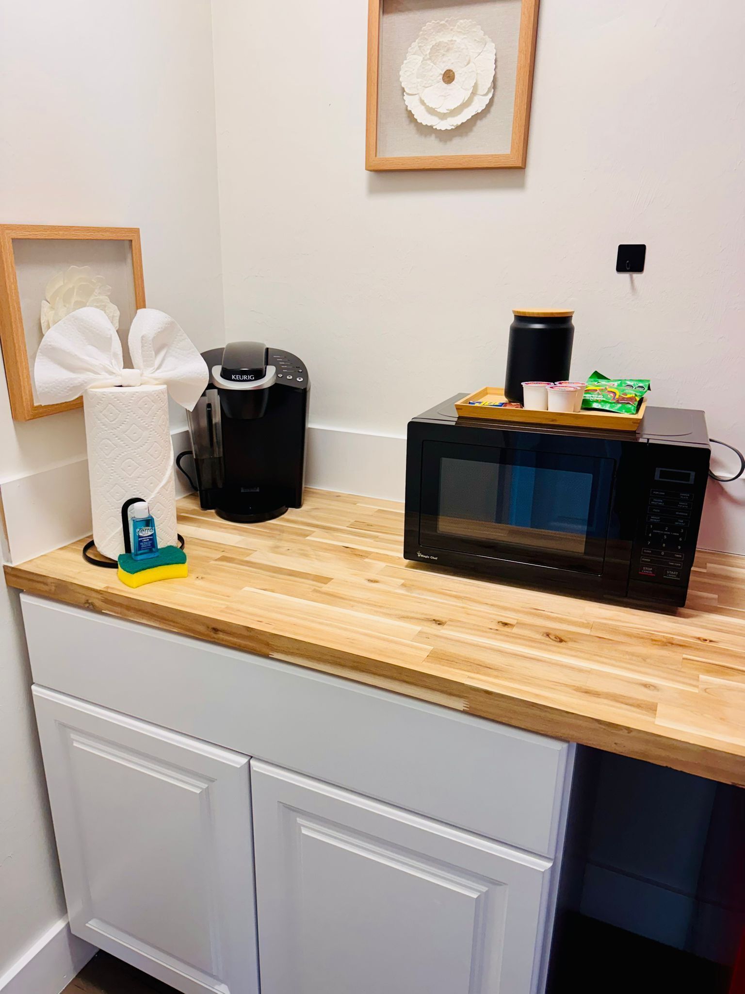 Een keuken met een houten aanrecht, een zwarte koffiezetapparaat, een magnetron en een decoratief object in een lijst aan de muur.