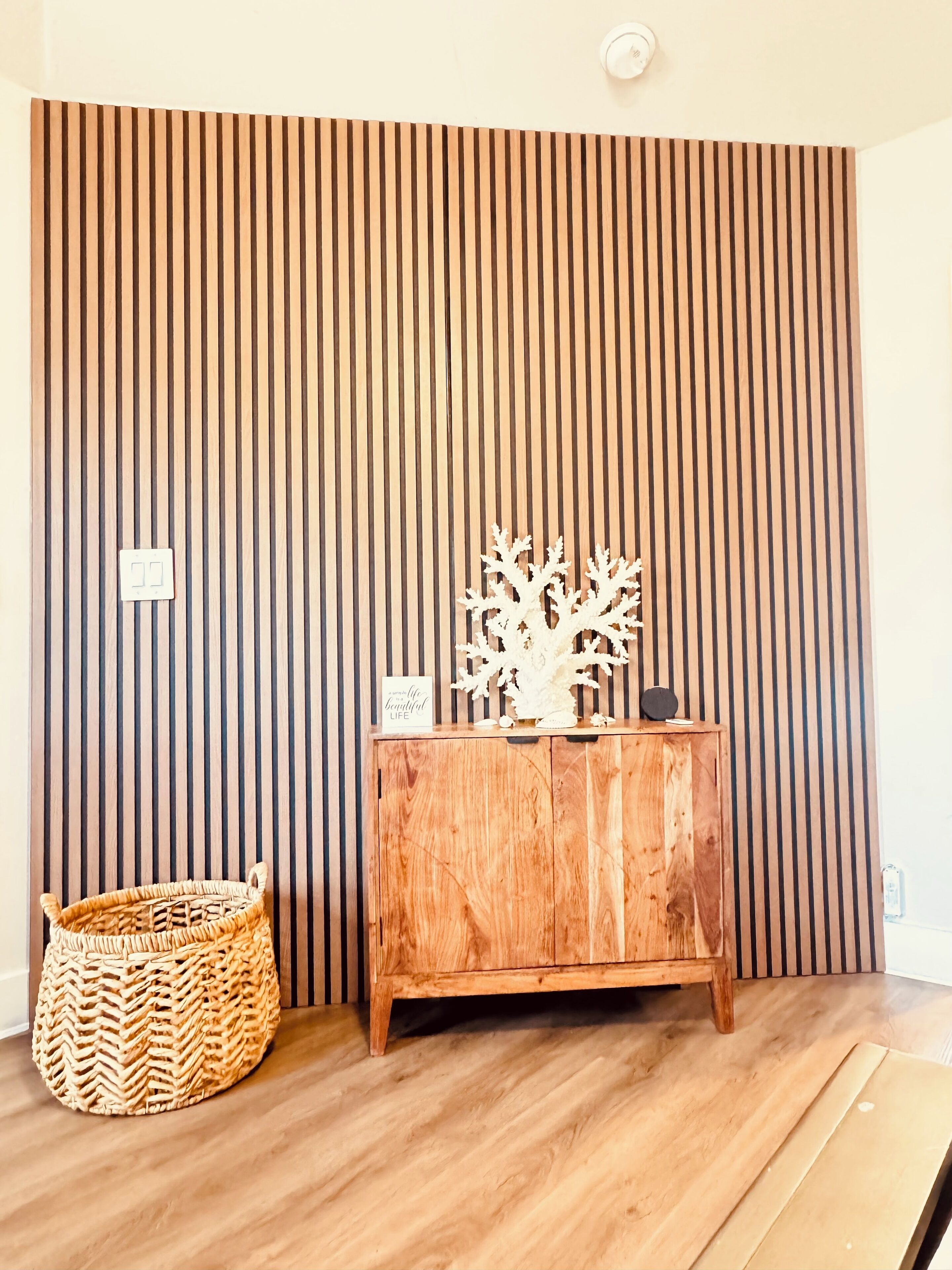 Une pièce avec un mur recouvert de lambris en bois, un meuble en bois orné d’objets décoratifs et un panier tressé posé sur le sol.