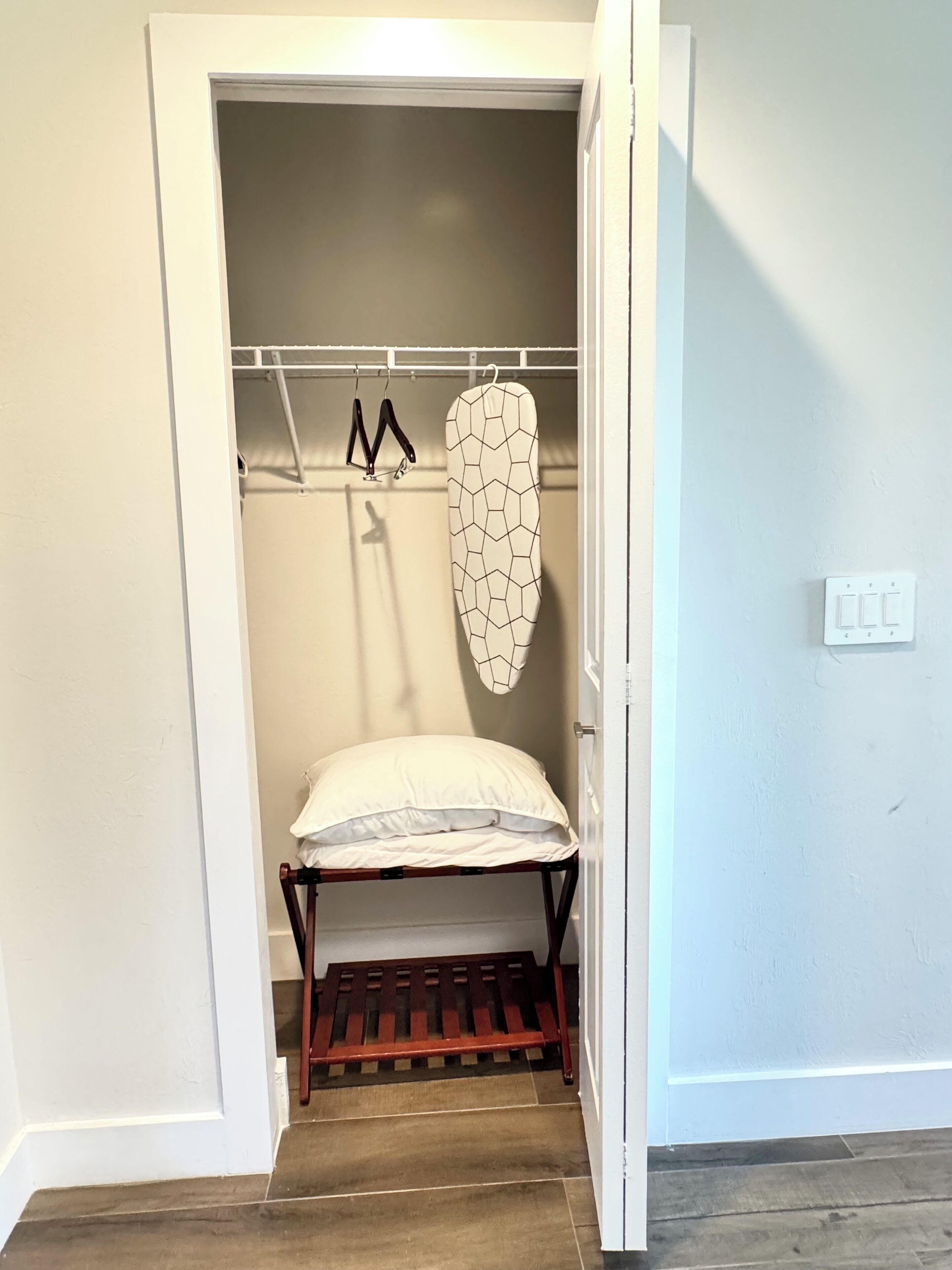 A small, enclosed closet with a wooden bench, a hanging iron board, and a coat rack.