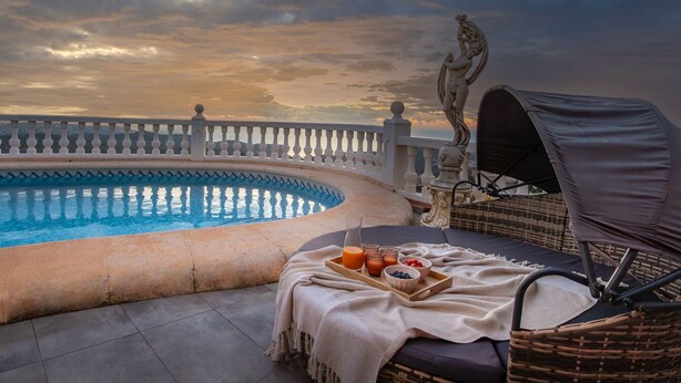 Zona junto a la piscina con una tumbona, una mesa con alimentos para el desayuno y una estatua.