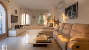 Una sala de estar moderna con un sofá color beige, una mesa de centro y un televisor. Hay ventanas con cortinas, una chimenea y un cuadro en la pared.