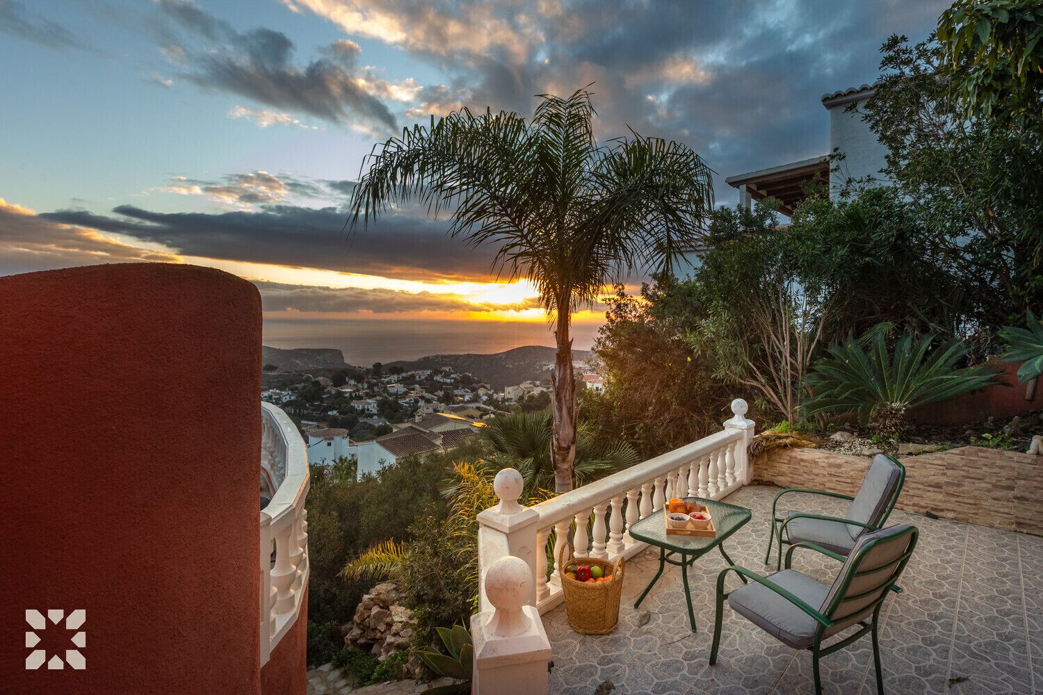 A terrace with a view of a coastal town at sunset, featuring a table with chairs, a small garden, and a decorative fence.