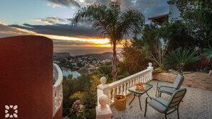 Una terraza con vistas a un pueblo costero al atardecer, con una mesa y sillas, un pequeño jardín y una valla decorativa.