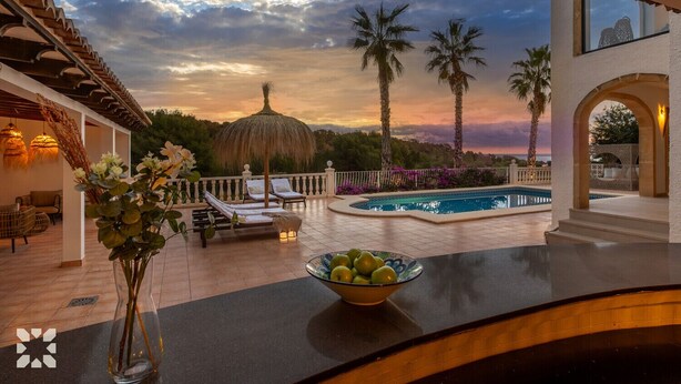 Un espace aménagé au bord de la piscine, avec des chaises longues, un parasol en chaume et une vue sur les palmiers et le coucher du soleil.