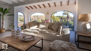 Un salon spacieux avec un canapé beige, une table basse et une vue sur une forêt, visible à travers de grandes fenêtres.