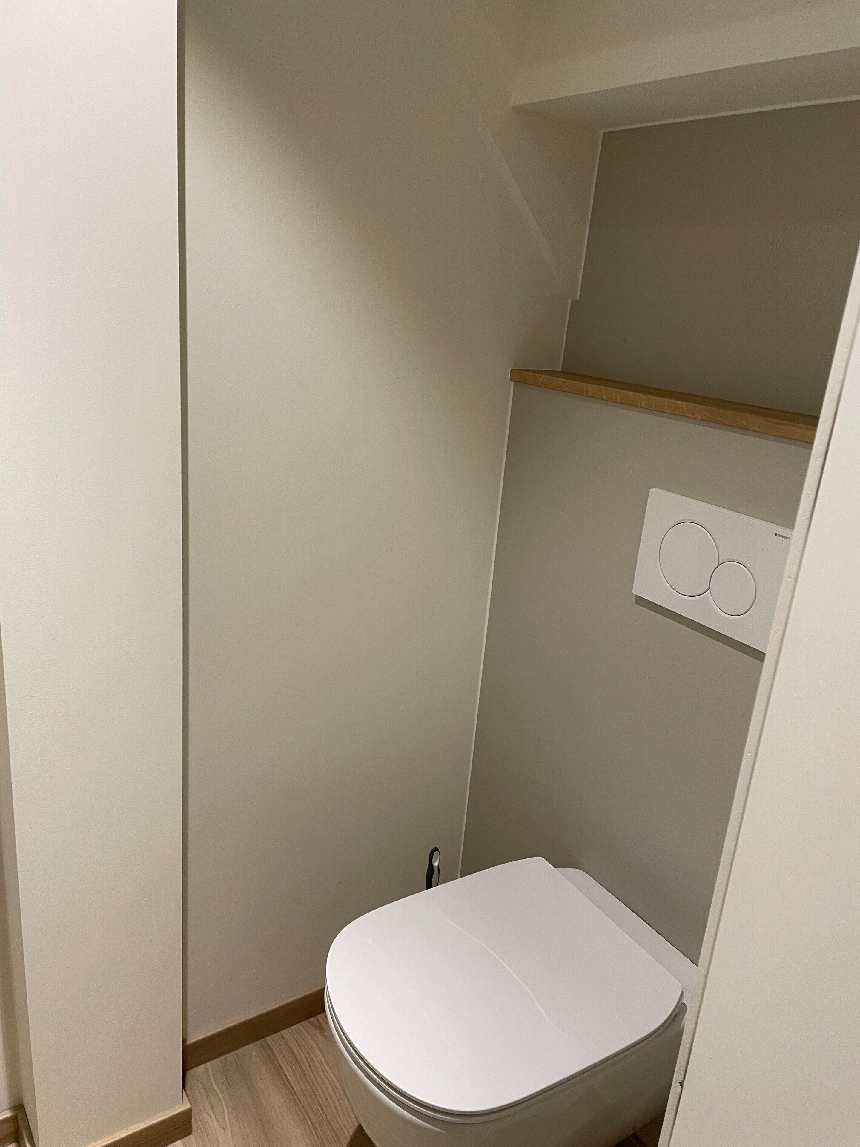 Un baño pequeño y funcional con inodoro blanco, estante de madera sobre el inodoro y aire acondicionado de pared.