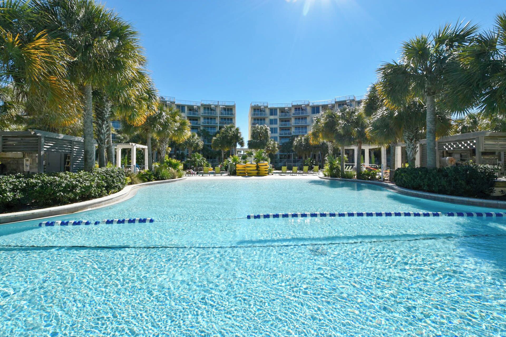 A large, clear swimming pool with blue lane dividers, surrounded by palm trees and a multi-story hotel building in the background.