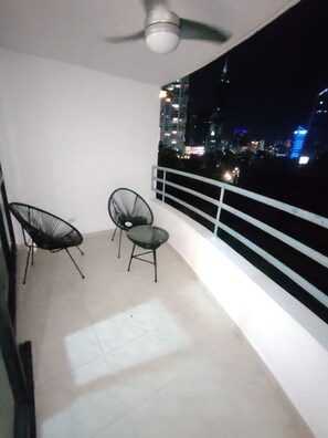Ein Balkon mit Deckenventilator, zwei Stühlen und einem Geländer mit Blick auf eine nächtliche Stadtlandschaft.