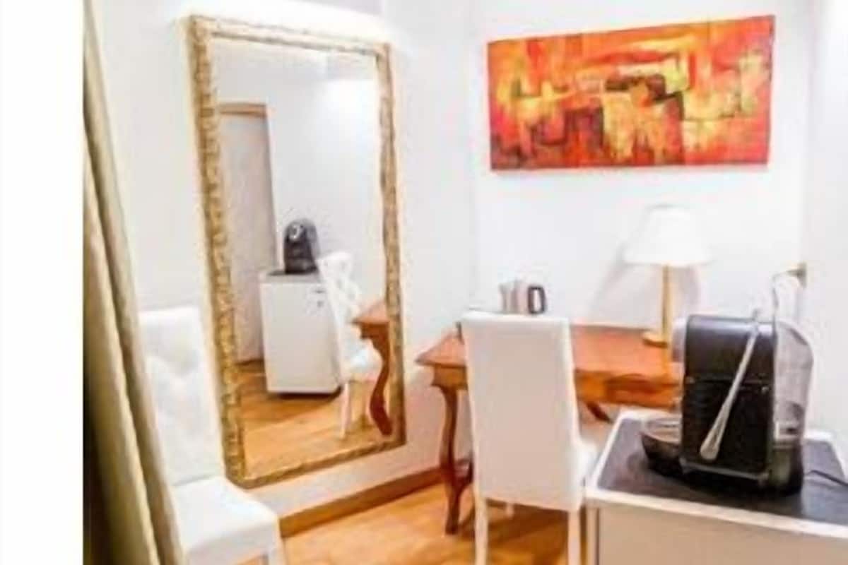 A compact room with a wooden floor, a white desk, a chair, a large mirror, and a painting on the wall.