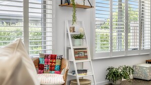 Una habitación luminosa con ventanas amplias, una escalera blanca con plantas, una silla de mimbre y una mesita con una planta.