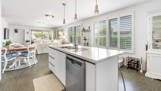 Una cocina moderna con una isla central, armarios blancos y lavavajillas. Varias ventanas con persianas, un área de comedor con mesa y sillas, e isla de cocina con fregadero y encimera.