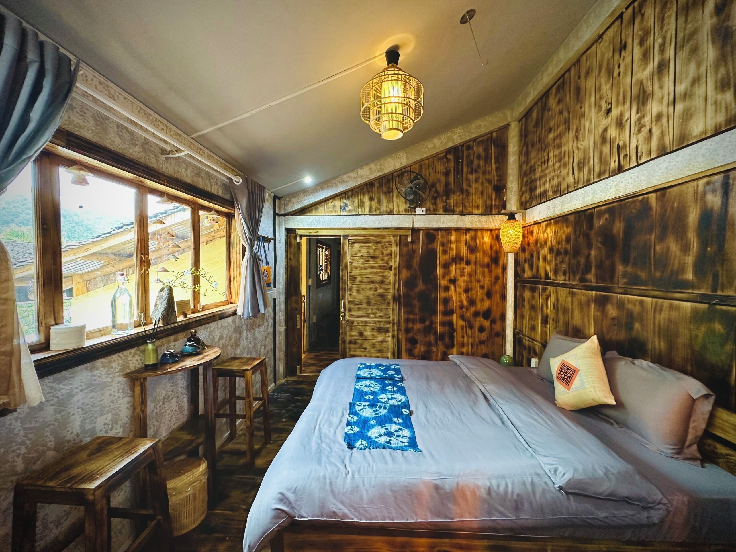 Une pièce avec un lit, des murs en bois, une fenêtre avec des rideaux, une petite table et un luminaire suspendu.