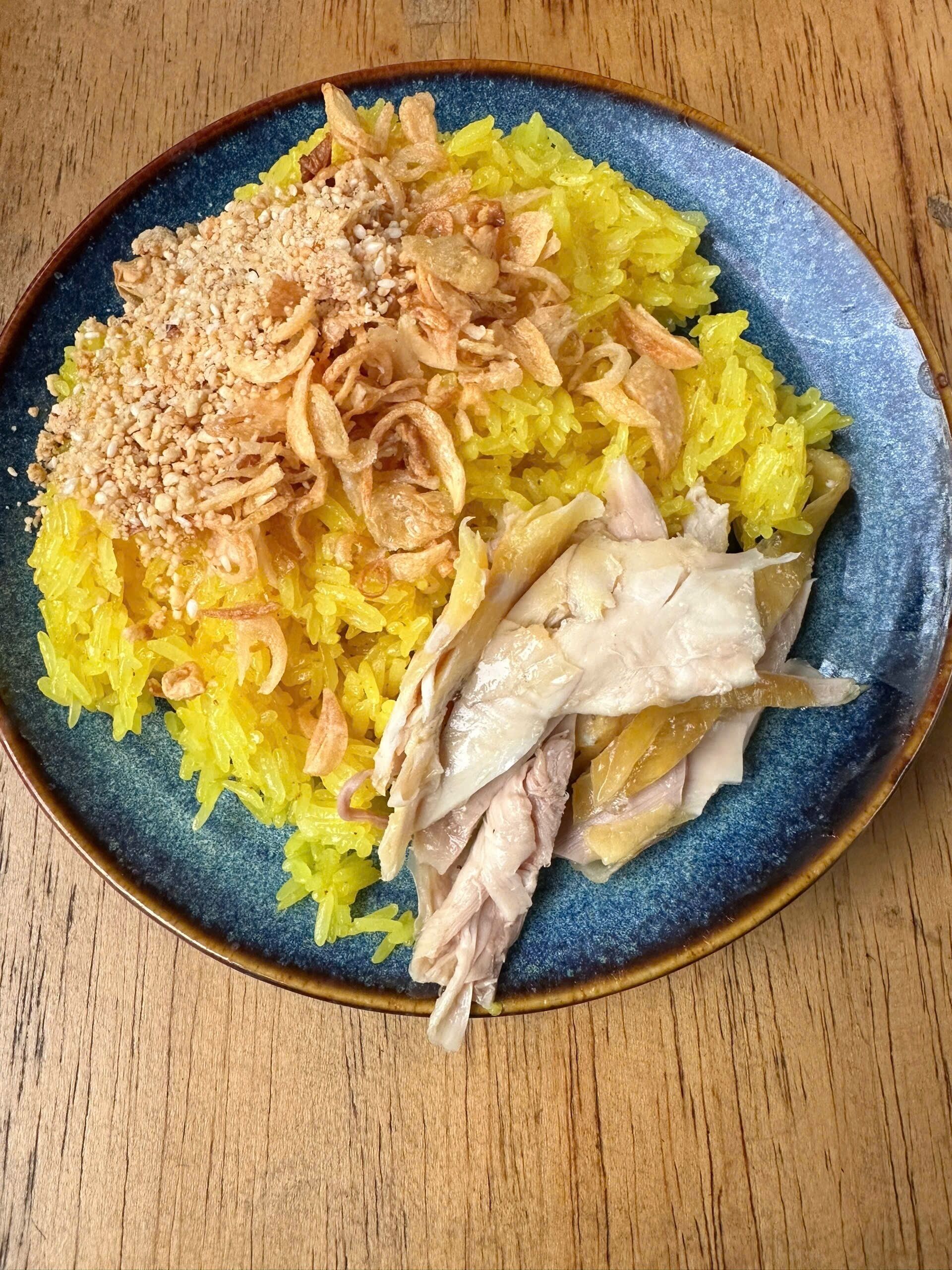A plate of yellow rice with fried onions and peanuts, and a piece of chicken on a blue plate.
