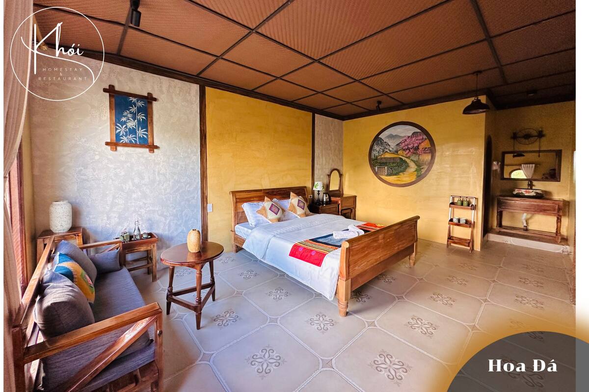 A cozy room with a bed, a small table, and a chair. There is a painting on the wall and a shelf with items on it.