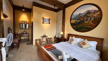 Quarto de hotel com cama grande, cabeceira de madeira, pintura de paisagem, pequena mesa com abajur e um ventilador.