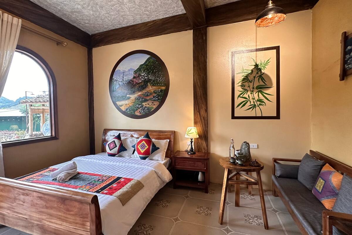 A bedroom with a wooden bed, a round painting, a bamboo plant, and a view of a building outside.