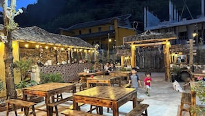 An outdoor dining area with rustic wooden tables and benches, surrounded by potted plants and a stone wall.